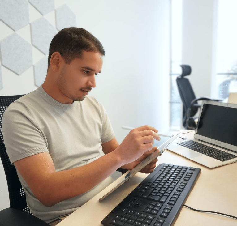 Werkstudent arbeitet konzentriert mit einem Tablet am Schreibtisch, neben Tastatur, Maus, Laptop und Notizblock. Er ist in der Vorbereitung seiner Bachelorarbeit.